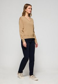 Beige v-udskæring sweater med lange ærmer, parret med mørkeblå bukser og lyse beige sneakers, præsenteret mod en neutral baggrund.