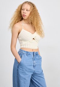 Top de punto color crema, corto, con parte delantera cruzada, tirantes finos y un patrón texturizado; combinado con unos pantalones vaqueros de mezclilla azul claro de tiro alto.