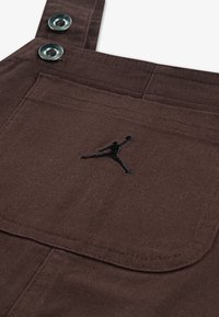 Pruunid puuvillased üldpusad metallnuppudega, millel on ees tasku. Taskul musta tikkimisega Jumpman logo. Tekstuuriga kangas.