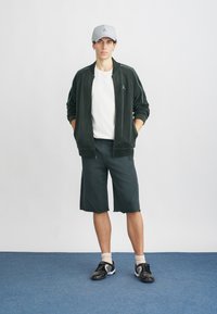 Casaco de treino de veludo verde escuro com detalhes azuis, t-shirt branca e calções a condizer. Boné cinza com logótipo e sapatilhas pretas de desporto.