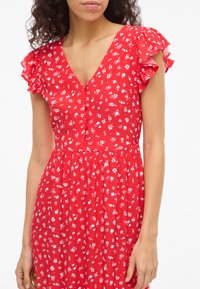 Donna che indossa un vestito rosso con stampa floreale bianca, scollo a V, davanti con bottoni e maniche corte arricciate su sfondo bianco.