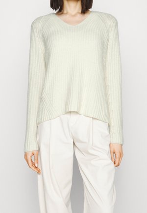 Frau trägt einen cremefarbenen, gerippten Strickpullover mit langen Ärmeln und weiße, plissierte, hoch taillierte Hosen vor einem schlichten Hintergrund.