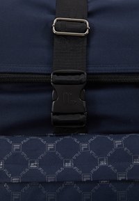 Modrá textilní taška s černým popruhem a plastovou sponou, zipovým uzávěrem a opakujícím se vzorem loga Fila v světlejší barvě.