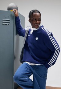 Μπλε σκούρο φούτερ με λευκές ρίγες στα μανίκια, με λογότυπο; συνδυασμένο με μπλε τζιν και γκρι πουλόβερ περασμένο στους ώμους.