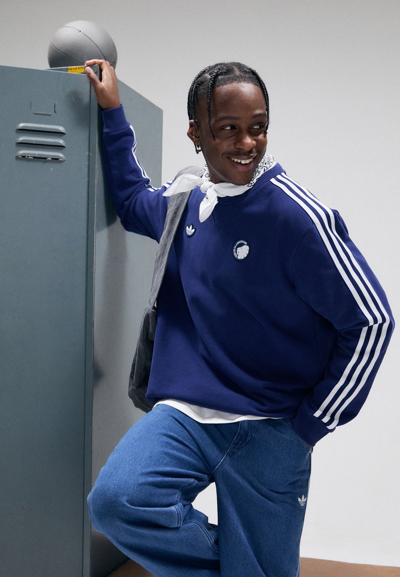 Μπλε σκούρο φούτερ με λευκές ρίγες στα μανίκια, με λογότυπο; συνδυασμένο με μπλε τζιν και γκρι πουλόβερ περασμένο στους ώμους.