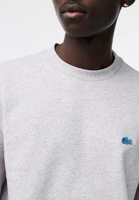 Sudadera gris claro con manchas multicolores, cuello redondo y logo bordado en el pecho. Tejido texturizado con un ajuste casual.
