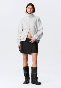 Szürke cipzáras pulóver magas gallérral, fekete átlapolt szoknyával és térdig érő fekete csizmával párosítva. Simas textúra, laza szabás, minimalista design.