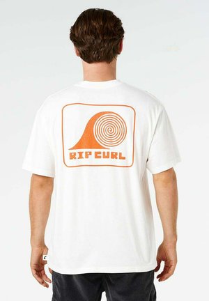 Mężczyzna o brązowych włosach stoi tyłem, ubrany w białą koszulkę z pomarańczowym logo spiralnej fali i napisem "RIP CURL" na plecach.