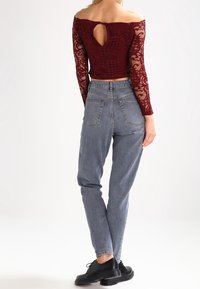 Žena oblečená v burgundském krajkovém topu bez ramínek s klíčovou dirkou na zádech, v šedých džínech vysokého pasu a černých šněrovacích botách, stojí s obratem k fotografovi.