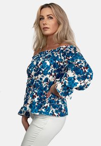Bluzka na jedno ramię w białym kolorze z niebieskim i burgundowym wzorem kwiatowym. Posiada falbaniasty dekolt oraz rękawy, a także rozkloszowany dół.