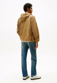 Chaqueta con capucha de color marrón claro, con puños y dobladillo elásticos, llevada sobre una camisa blanca y jeans azules, combinada con zapatillas blancas.