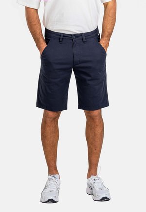Male model wearing navy blue knee-length shorts with hands in pockets, paired with a white t-shirt and white sneakers, standing on white background.