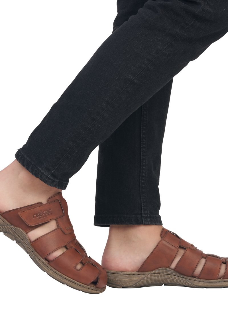 Brown leather sandals with open design, featuring cut-outs and a textured sole. Paired with black denim pants, showcasing casual styling.