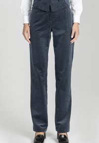 Kobieta w ciemnoniebieskich sztruksowych spodniach, białej koszuli i czarnych butach, stojąca na jednolitym jasnoszarym tle.