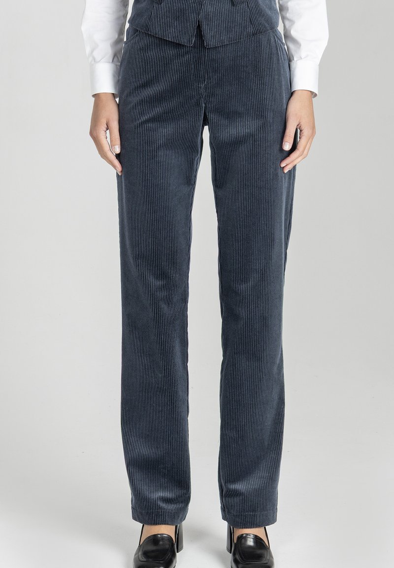 Kobieta w ciemnoniebieskich sztruksowych spodniach, białej koszuli i czarnych butach, stojąca na jednolitym jasnoszarym tle.