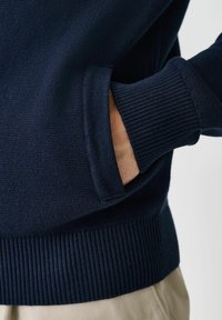 Navy blue knitted sweater with ribbed cuffs and hem, featuring a side pocket and a smooth, textured finish.