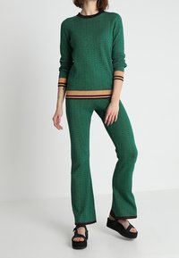 Grüner Fischgrätenmuster-Strickpullover und ausgestellte Hose mit orangefarbenen Akzenten. Sandalen mit dickem schwarzem Sohle. Weiche Textur, lässige Passform.