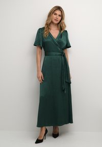 Rochie verde dintr-un material asemănător satinului, cu mâneci scurte tip balon, o legare la talie și o fustă lungă, fluidă. Inclusiv tocuri negre.