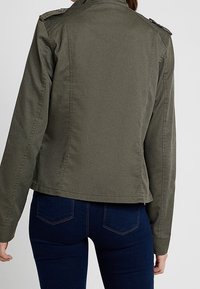 Kvinna i olivgrön militarinspirerad jacka och mörkbl�å jeans, sedd bakifrån mot en enkel ljus bakgrund.