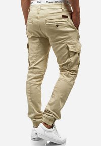 Pantalons cargo beige avec poignets élastiques, plusieurs poches et une taille Calvin Klein. Le matériau semble être du coton avec une texture lisse.