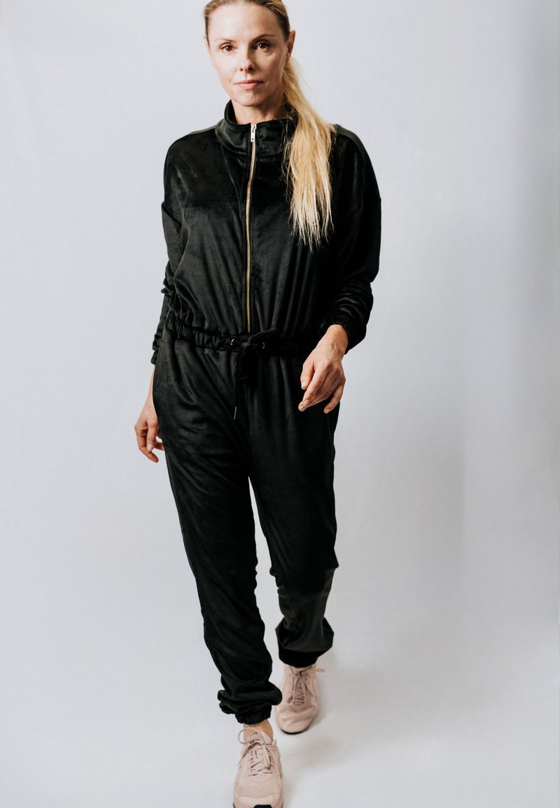 Naine kõndimas otse musta tõmblukuga kombinesooni ja heleroosade tossudega halli tausta ees.