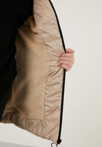 Beige isoleret jakke med en blød, fluffy inderside. Har et quiltet mønster og en sort lynlås langs siden. Hånd der holder lynlåsen.