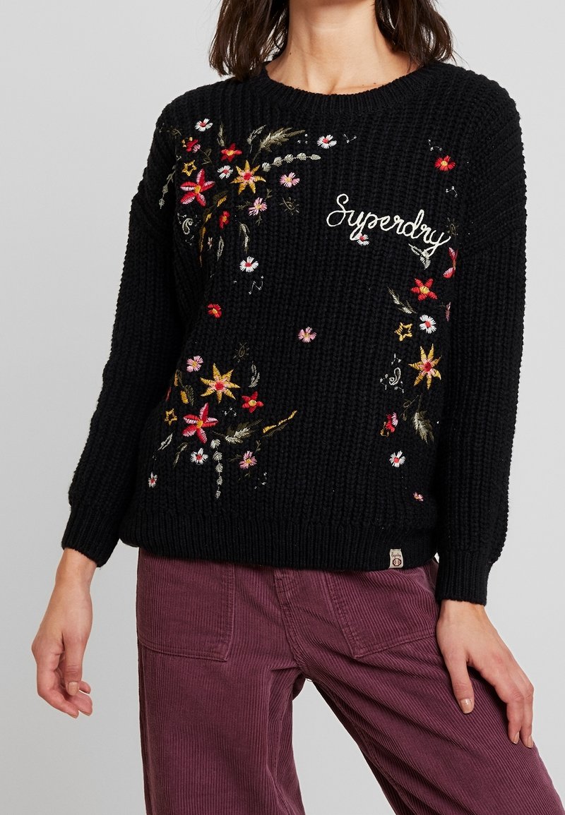 Pull en maille noir avec broderie florale colorée et texte "Superdry", associé à un pantalon en velours côtelé violet taille haute.