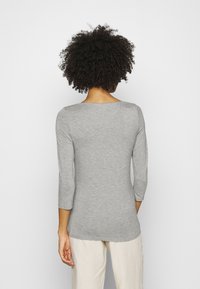 Nainen, jolla on kihara hius ja päällään vaalea harmaa pitkähihainen paita ja vaaleat housut, kääntyy poispäin yksinkertaista valkoista taustaa vasten.