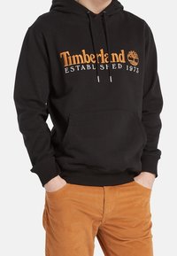 Uomo che indossa una felpa nera Timberland con logo e pantaloni in velluto a coste marroni, con le mani nelle tasche, su uno sfondo semplice.