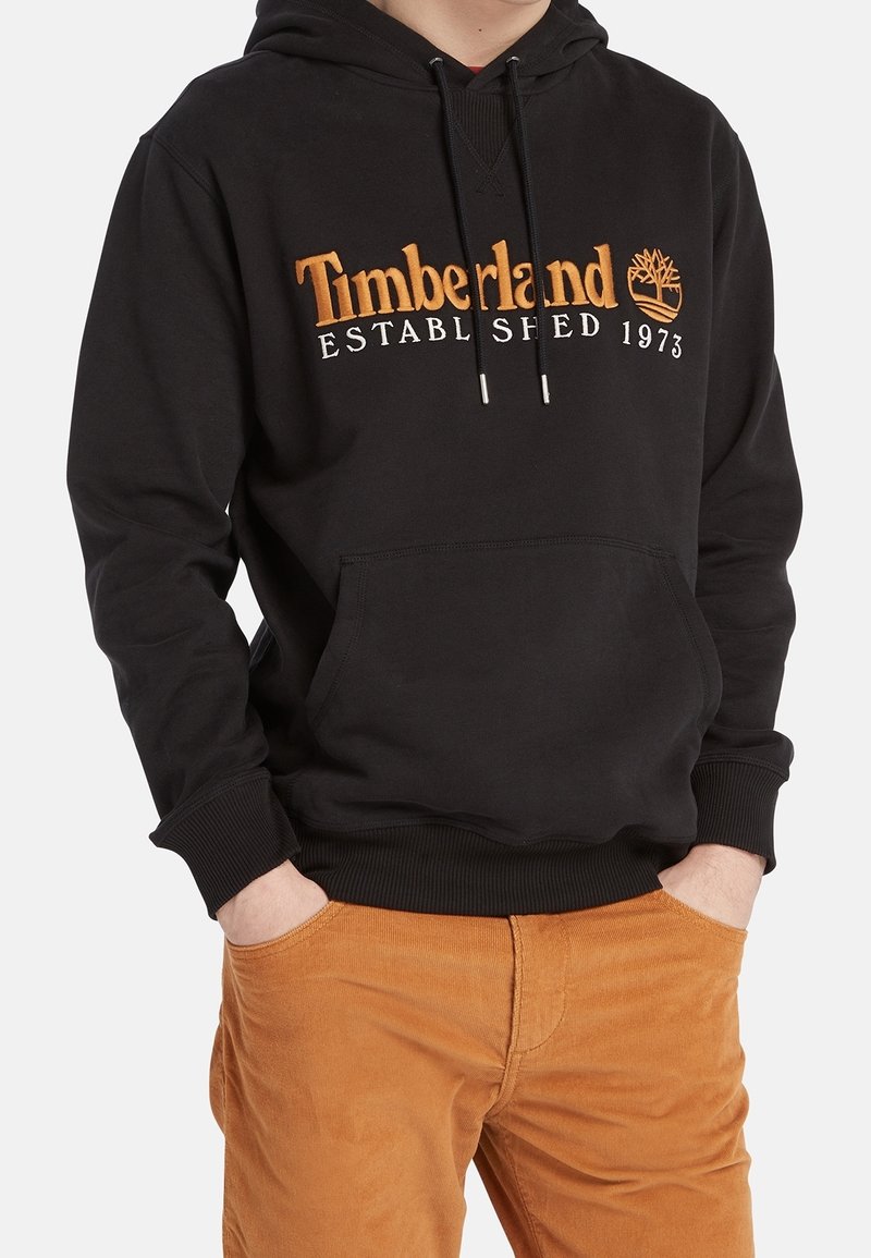 Uomo che indossa una felpa nera Timberland con logo e pantaloni in velluto a coste marroni, con le mani nelle tasche, su uno sfondo semplice.