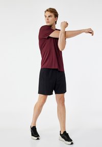 Bordeauxfarbenes kurzes Sportoberteil und schwarze Shorts. Das Modell streckt einen Arm aus. Die Schuhe sind schwarz mit weißen Sohlen. Einfaches und funktionales Design.