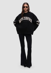 Schwarzer oversized Pullover mit cremefarbenem Aufdruck "WRSTBHVR", fransigem Saum, kombiniert mit schwarzen gerippten Schlaghosen und spitzen Schuhen.