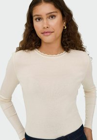 Langermet kremfarget topp med rund hals og bølgete blonderkant. Myk tekstur, tettsittende passform og uten synlige mønstre eller trykk.