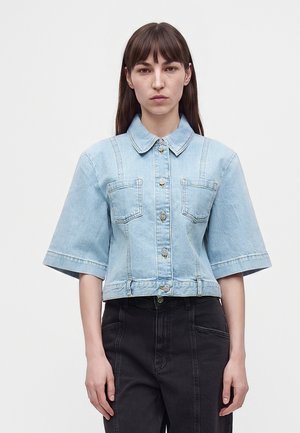 Femme aux cheveux bruns et raides porte une veste en jean bleu clair à manches courtes avec des boutons argentés et un jean noir taille haute sur fond blanc.