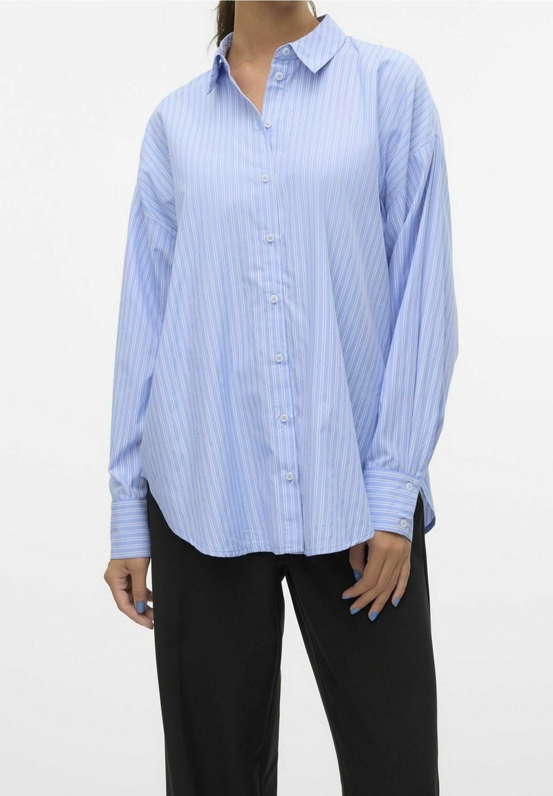 Femme portant une chemise ample à rayures bleu clair et blanc avec des boutons et des manches longues, et un pantalon noir, debout devant un fond blanc uni.