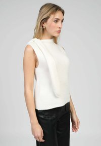 Femme aux longs cheveux blonds portant un pull sans manches blanc et un pantalon noir, tournée vers la droite sur un fond gris uni.