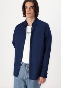 Giovane uomo con capelli alla lunghezza delle spalle che indossa una camicia blu scuro sbottonata sopra una t-shirt bianca con grafica e jeans chiari su sfondo bianco.