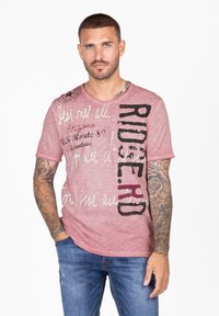 Camiseta rosa de acabado texturizado con texto gráfico en blanco y negro. Mangas cortas y escote redondo. Detalle de brazo tatuado.