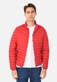 Giacca rossa imbottita con cuciture orizzontali, zip frontale e colletto rialzato. Indossata sopra una maglietta bianca con jeans blue. Materiale leggero.
