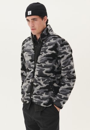 Chaqueta de forro polar con cremallera en patrón de camuflaje negro y gris, que cuenta con un alto cuello, dos bolsillos laterales y un acabado suave y texturizado.