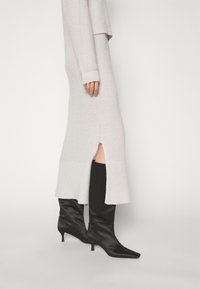 Robe grise en tricot avec une fente latérale, associée à des bottes noires montantes à bout pointu en matériau brillant. Présente une texture côtelée et des manches longues.
