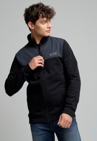 Giacca nera con zip e pannelli grigi sulle spalle; presenta un collo alto, polsini e orlo a costine, e un logo discreto sul petto.