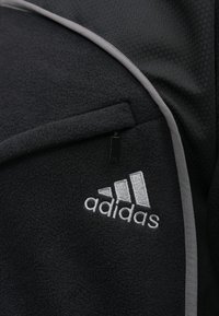 Melnas vilnas jaka ar pelēkiem akcentiem, kurai ir strukturēta šūnu forma, ar rāvējslēdzēja kabatu un izteiktu baltu izšūtu logotipu.