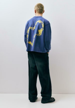 Personne aux cheveux blonds courts portant un sweat-shirt bleu marine avec un grand point d'interrogation jaune dans le dos, un pantalon noir et des chaussures noires, debout face à un mur.