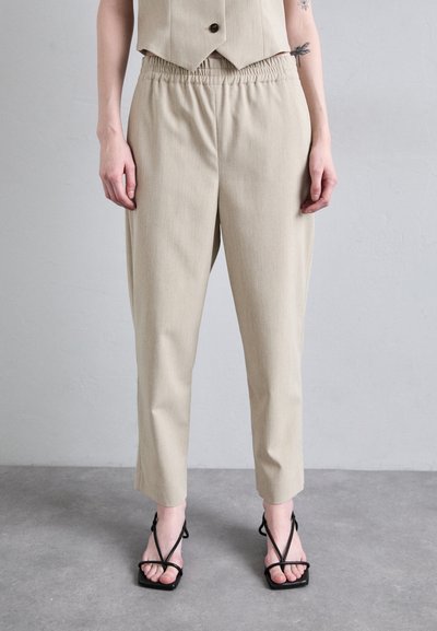 Pantalones beige ajustados con un corte relajado y cinturilla elástica, con un tejido texturizado. Combinados con sandalias negras de tiras.