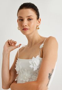 Top blanco con bordado floral y tirantes finos, con flores de encaje texturizadas y un diseño ajustado. La modelo tiene un tatuaje en el brazo.
