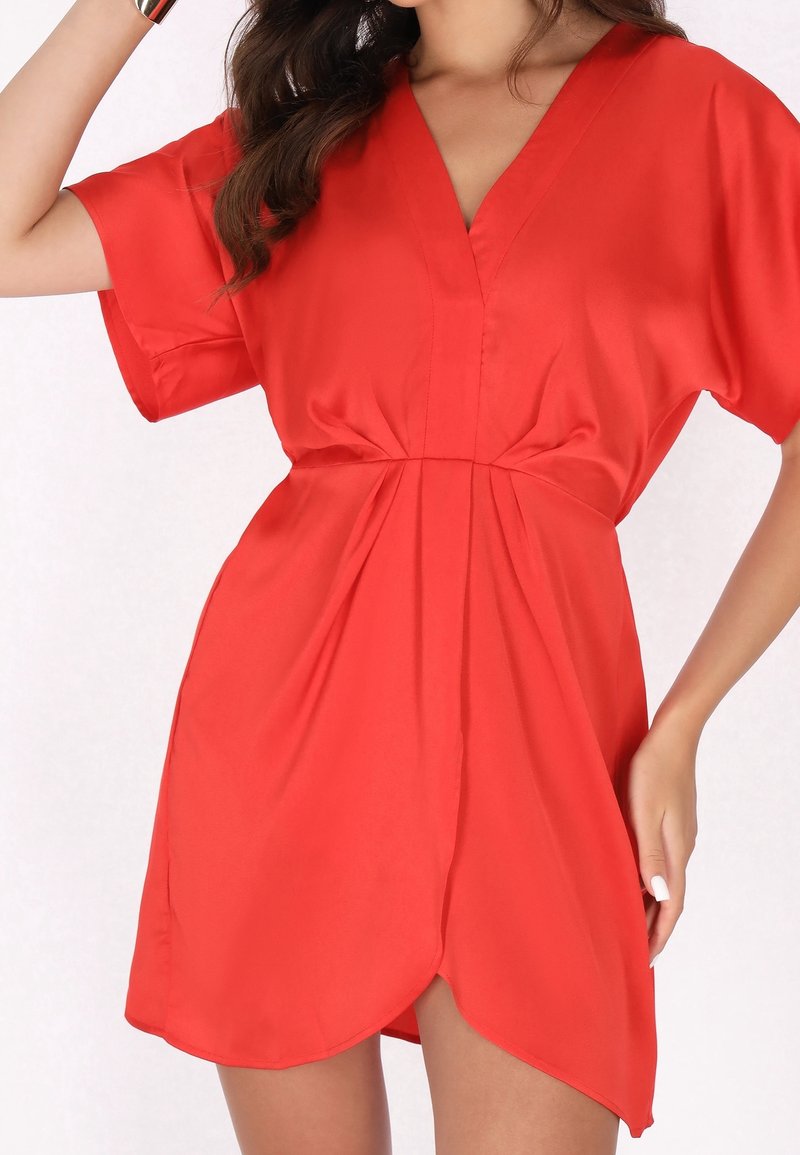 Femme portant une robe portefeuille rouge vif à manches courtes, avec un décolleté en V et une taille froncée, posant avec une main près de ses cheveux.
