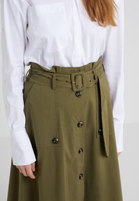 Gonna verde oliva con una cintura larga, dotata di grandi bottoni neri e due tasche frontali. Abbinata a una camicia bianca con bottoni.