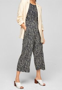 Marinblå overall med prickigt mönster i vitt och grönt, med cinchad midja, korta vida ben, och parat med en krämfärgad blazer och vita halkskor.