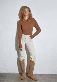 Brązowy sweter z prążkowanym wzorem i długim rękawem, kremowe dżinsy o wysokim stanie oraz beżowe kowbojki haftowane, z ostro zakończonym noskiem i stożkowym obcasem.